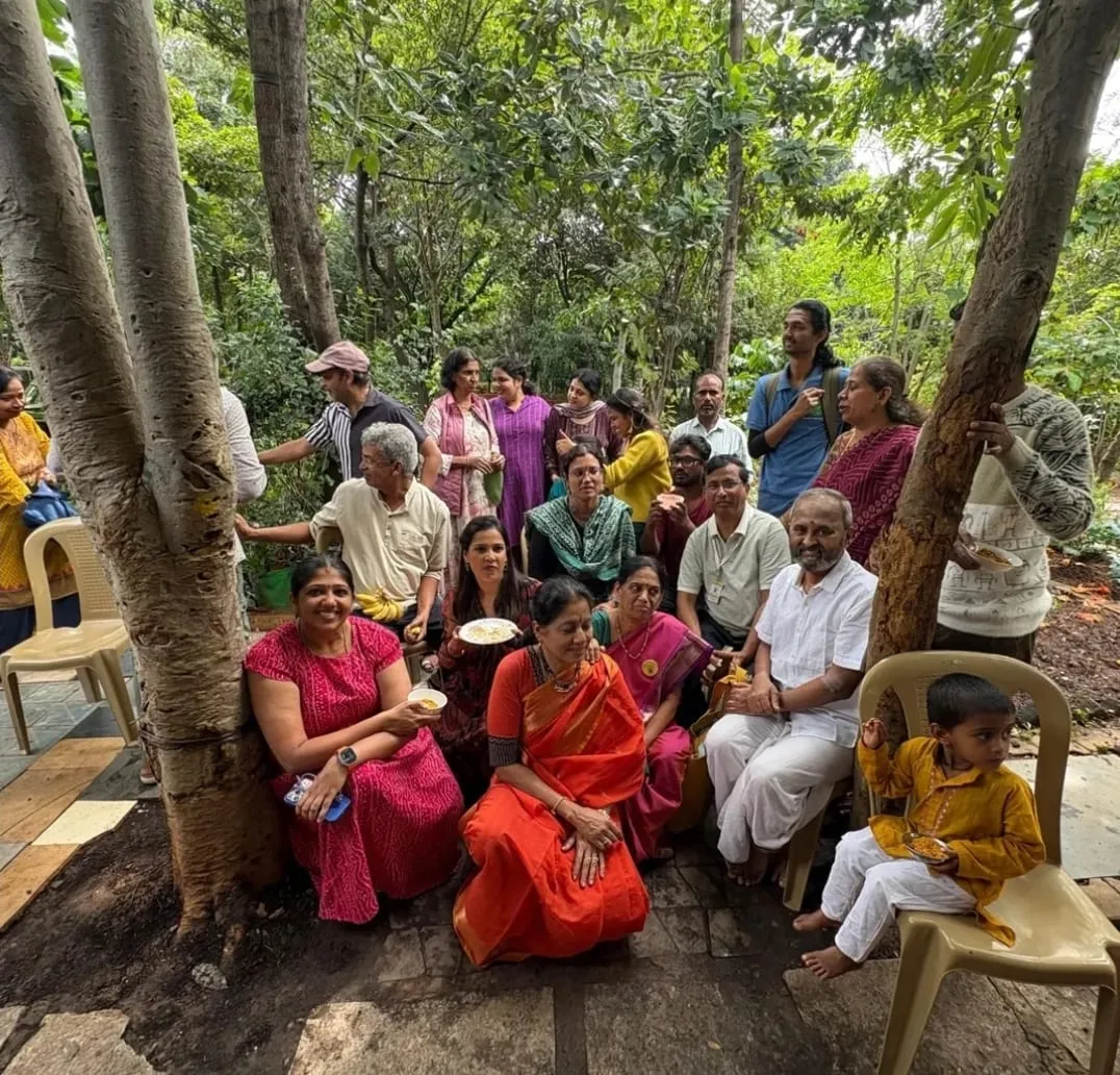 Volunteers and visitors of all ages gathered under trees in a leafy garden, some holding plates of food