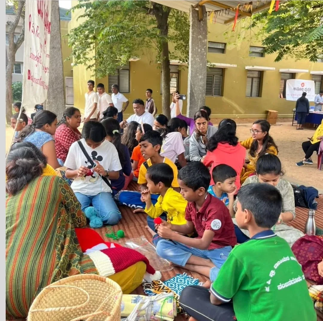 Children and adults sitting on mats under trees learning to crochet with balls of wool at an outdoor santhe