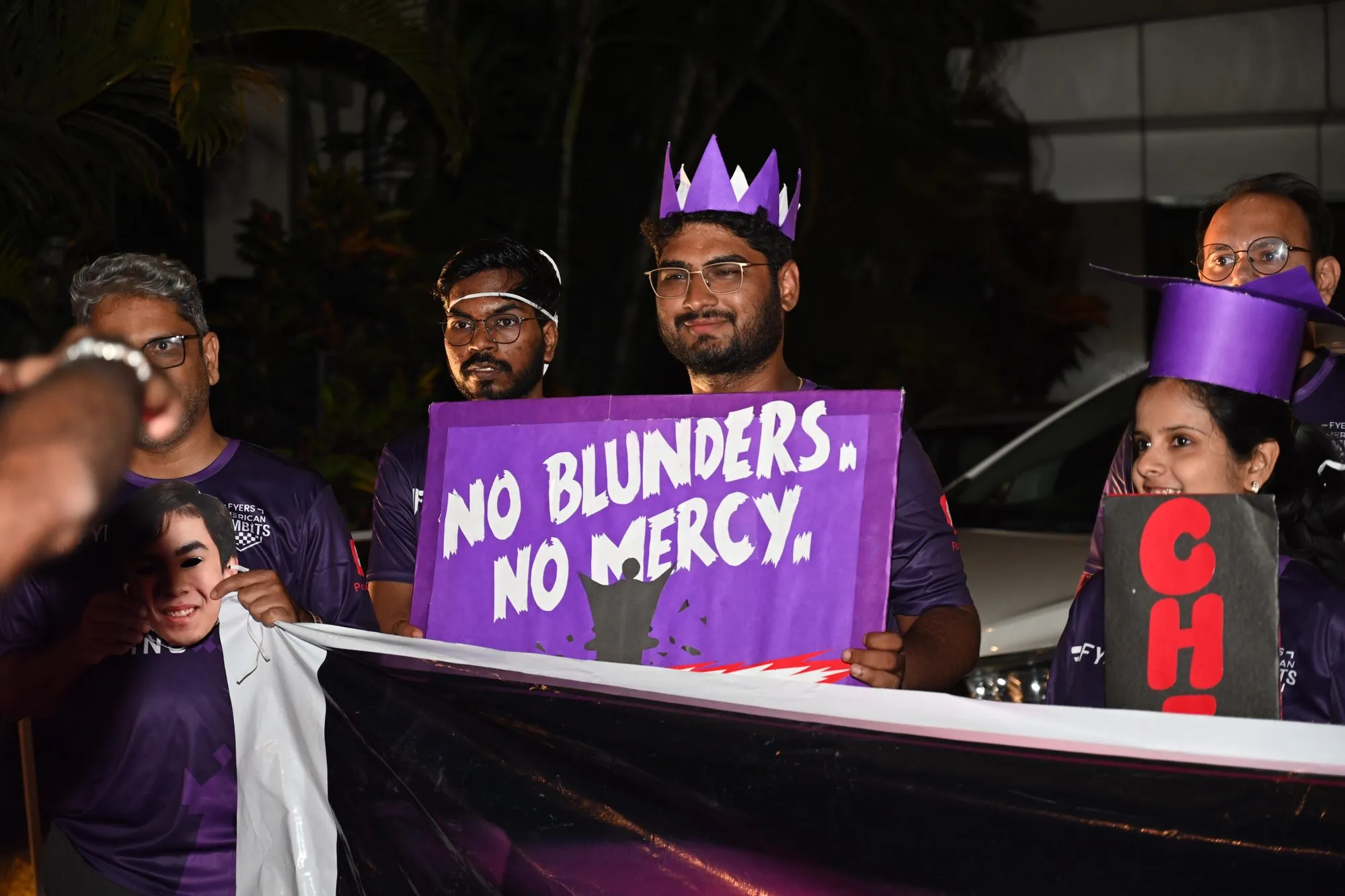 A volunteer in a paper crown holding a No blunders, no mercy placard at night