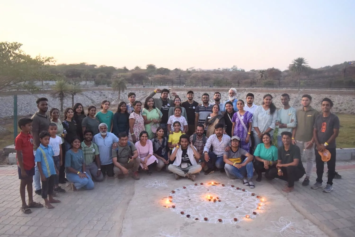 Volunteers, organisers and children pose for a group photo at Srigandhadakavalu lake at dusk, around a rangoli ringed with lit diyas