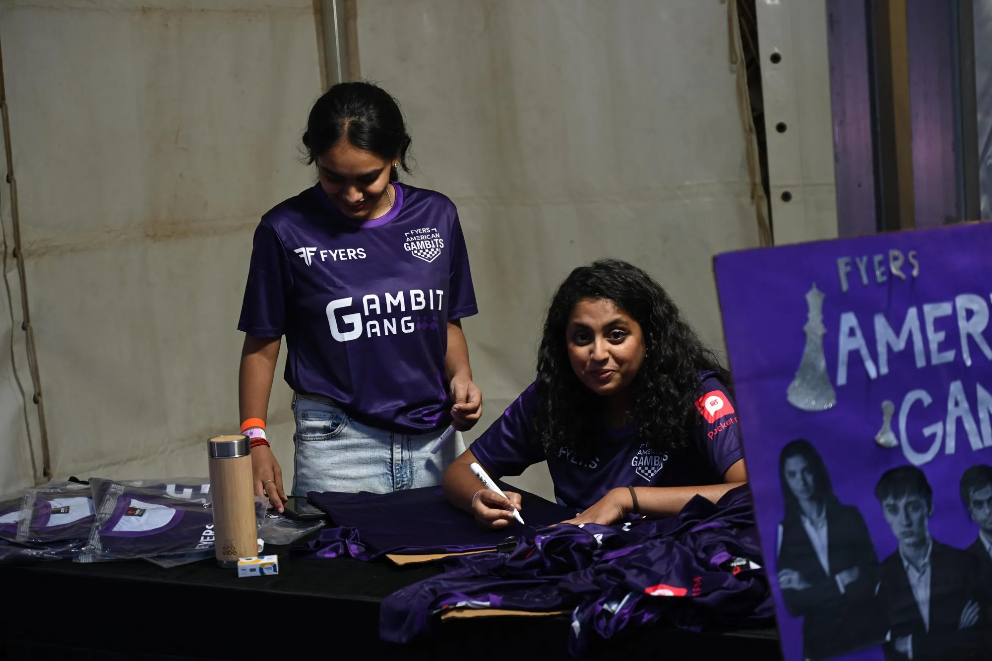 Two volunteers at the merch table, one writing on a jersey